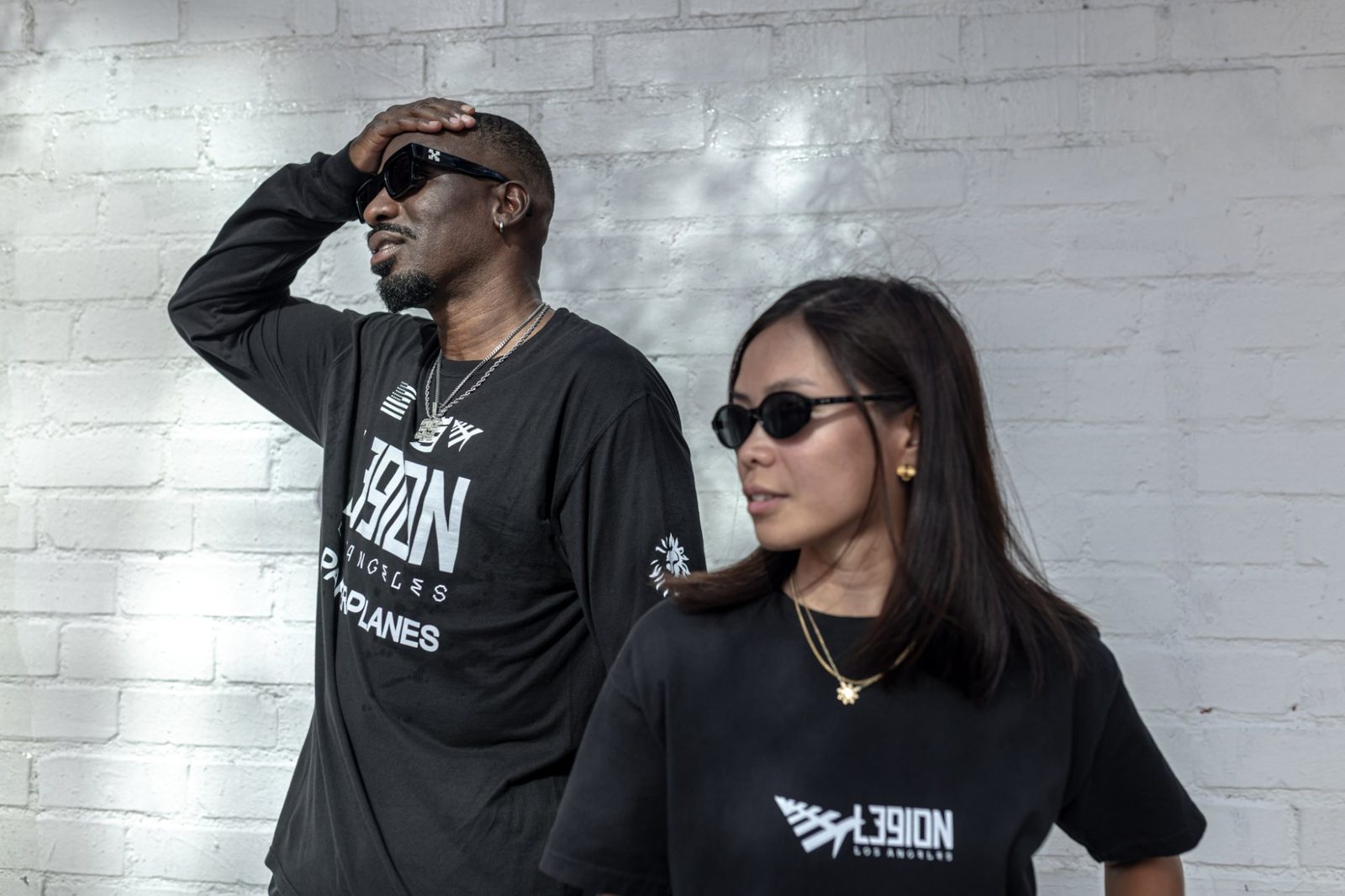 Two people in black L39ION Los Angeles Paper Planes tees against a white brick wall